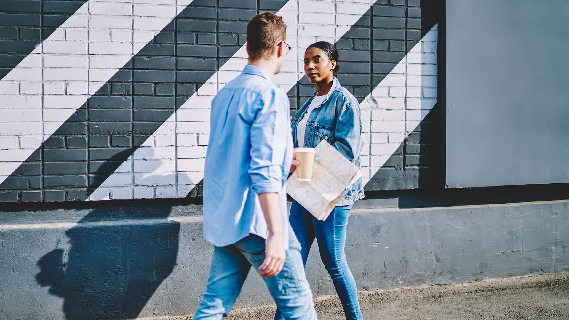 Man and woman dressed in light blue jeans making eye contact as they pass each other on the road while walking.